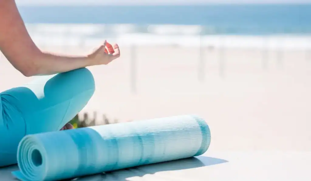 woman in blue sitting on beach with yoga mat